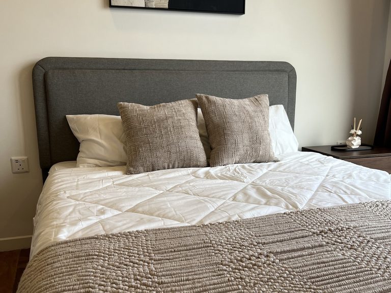 Close view of the bed with white quilted bedding, a beige textured throw and two cushions